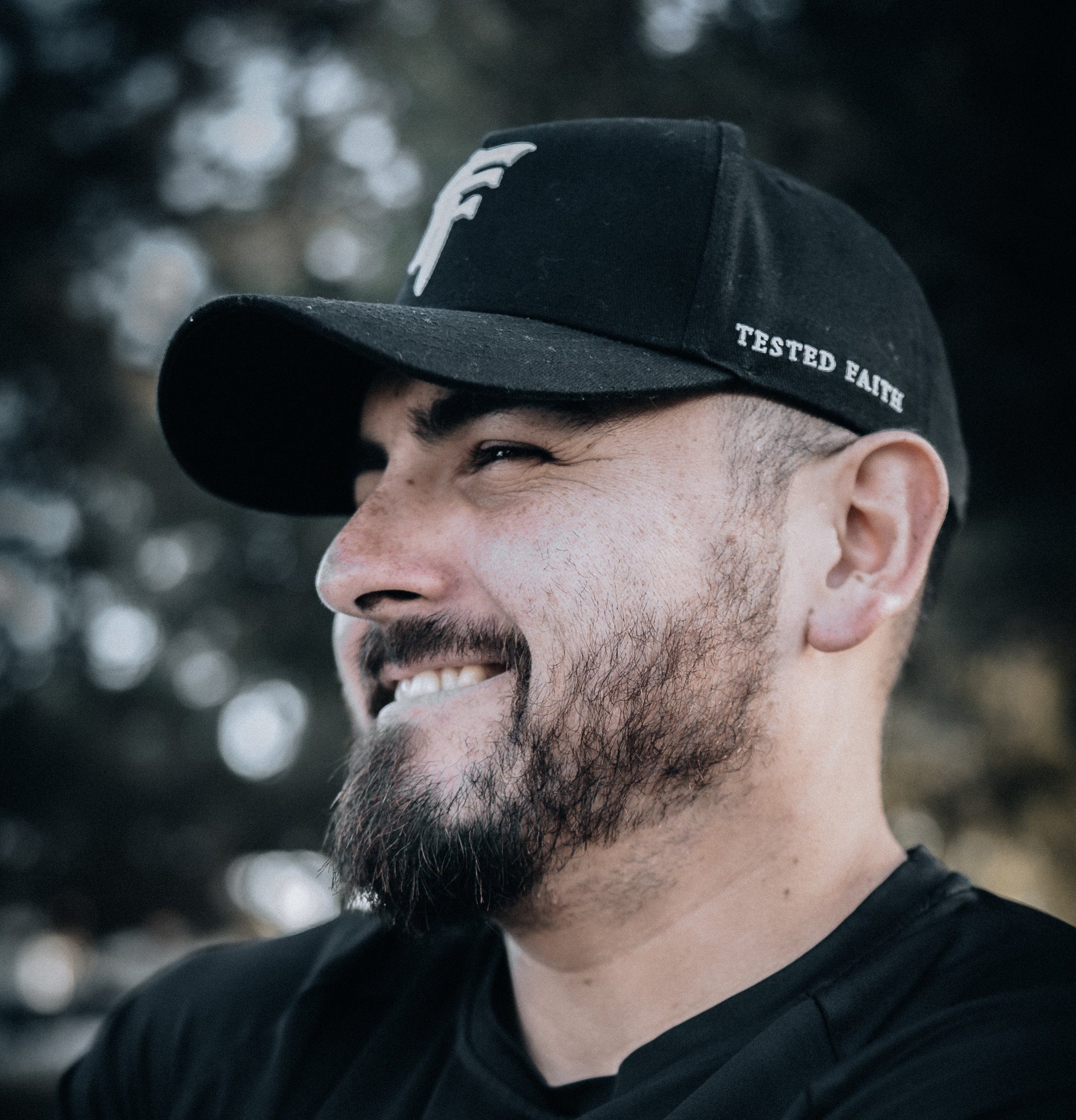 Man wearing a black cap and t-shirt with a logo, standing against a blurred natural background.